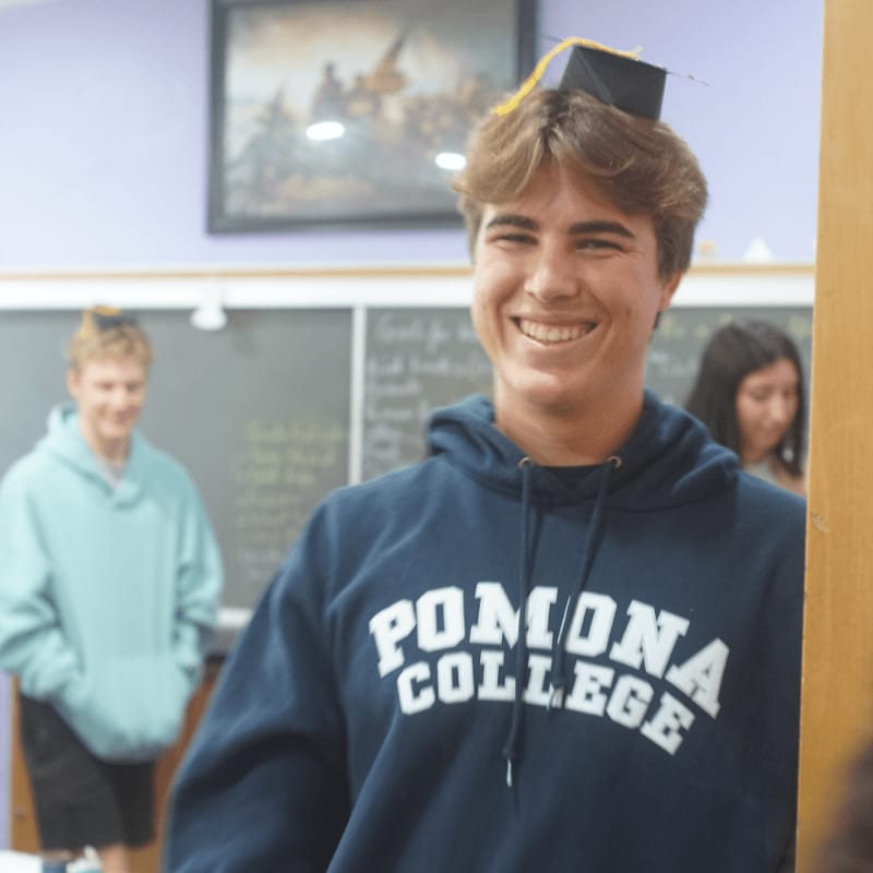 A smiling young man wearing a Pomona College hoodie and a small graduation cap sits in the foreground, with other students and a chalkboard in the background.