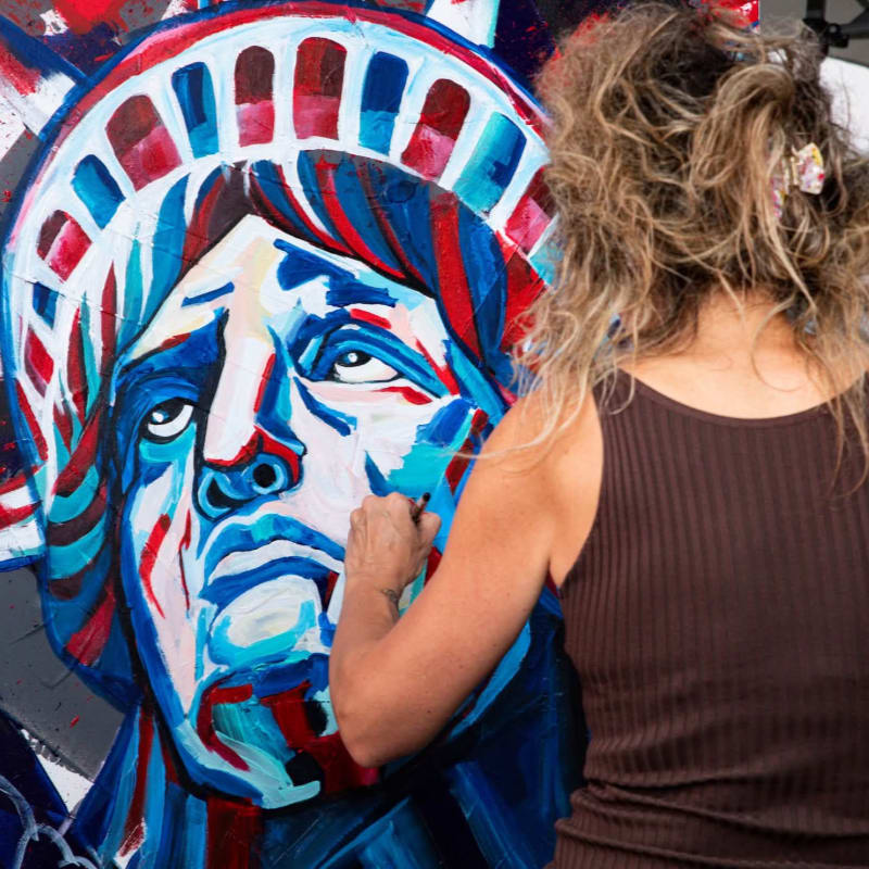 A woman with curly hair paints a colorful, stylized portrait of the Statue of Liberty on a large canvas.