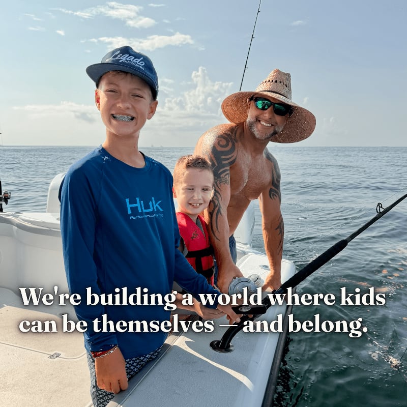 A man and two young boys smile on a boat against a backdrop of a calm ocean and a cloudy sky.