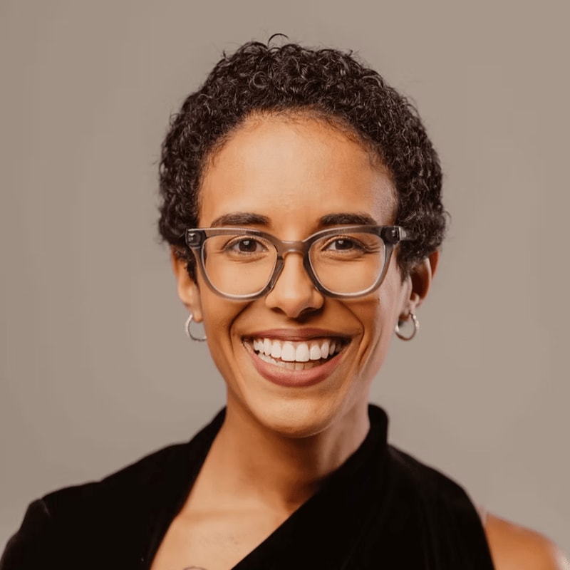 A smiling woman with short curly hair and glasses stands in the foreground against a plain, neutral grey background.