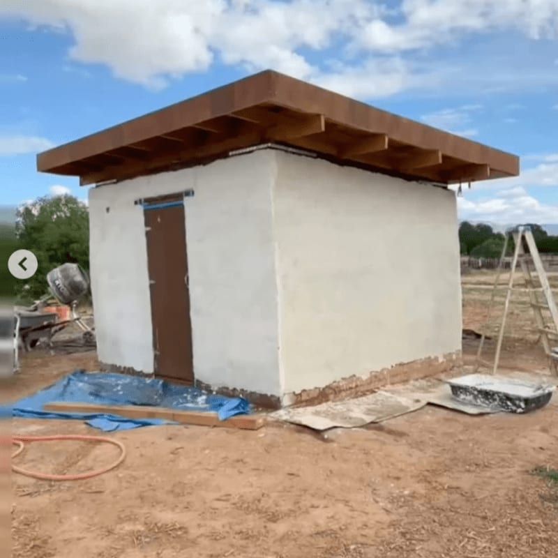 A small, white plastered building with a brown wooden roof stands on a dirt lot under a cloudy blue sky.