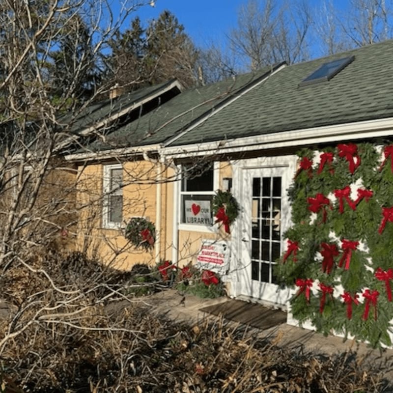 A yellow building decorated with Christmas wreaths and red bows stands behind a foreground of dormant, brown brush under a clear blue sky.