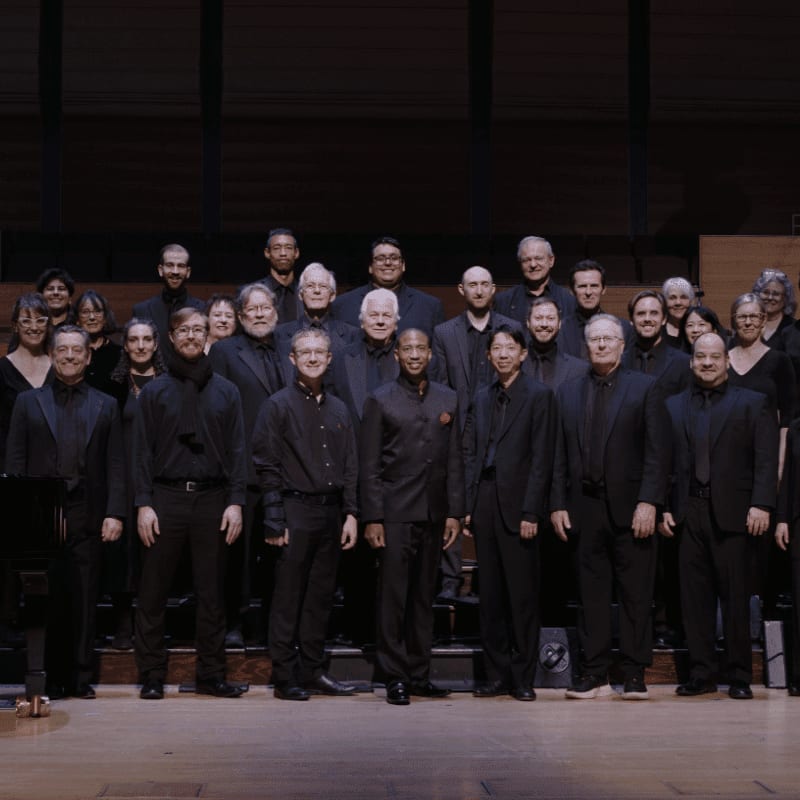 A choir of men and women in black attire stands on a stage in front of a dark, tiered background, with a piano visible on the left side of the foreground.