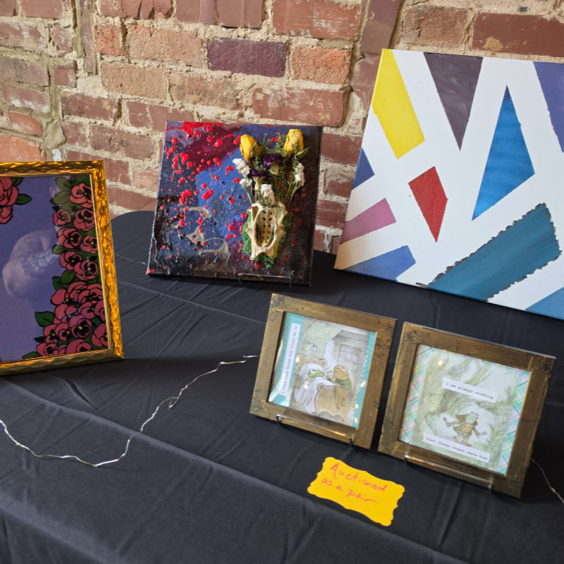 Various pieces of framed art sit on a black tablecloth in front of a red brick wall.