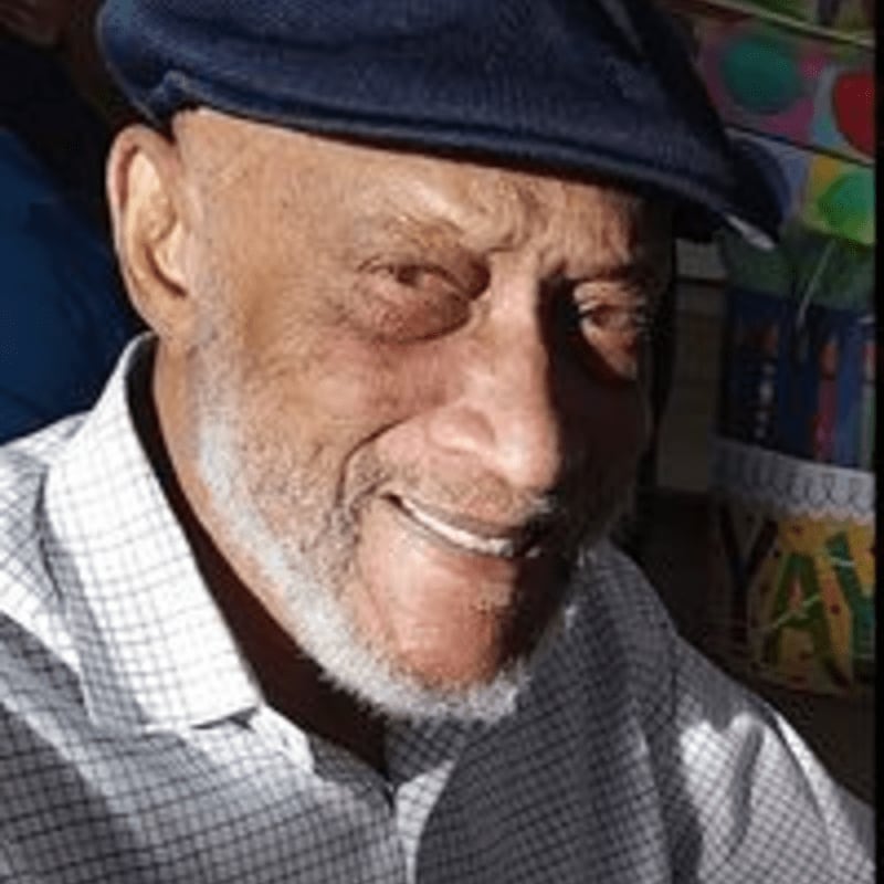 An elderly man wearing a dark blue cap and a checkered shirt smiles in the foreground against a colorful, blurred background.