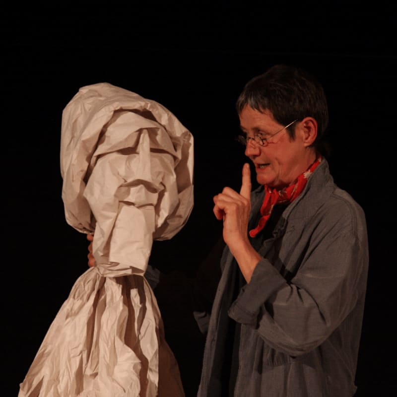 A woman with glasses and a red scarf gestures with her finger toward a large, crumpled paper figure against a black background.