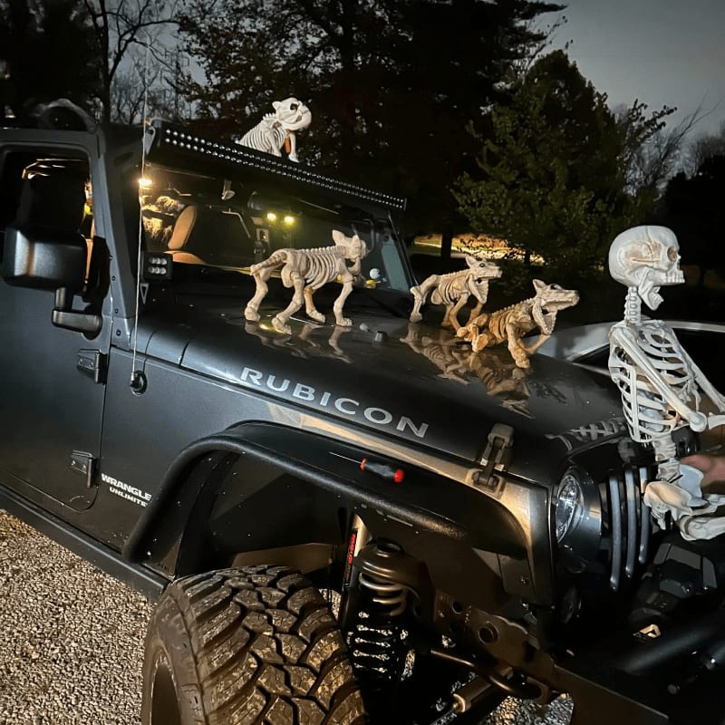 Several skeletal figures are perched on a dark grey Jeep Rubicon parked on a gravel surface against a backdrop of dark trees and a twilight sky.