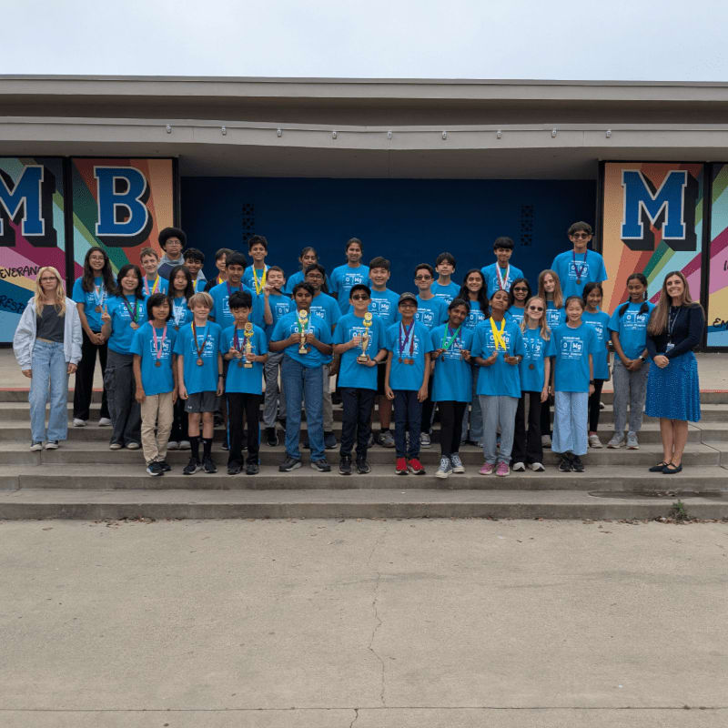A group of students in blue shirts holding trophies stands on concrete steps in front of a building with large letter signage.
