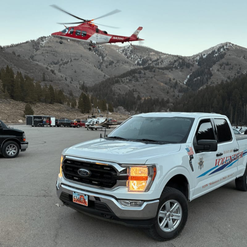 A white Ford pickup truck sits in a parking lot with a red and white helicopter flying over a mountainous landscape in the background.