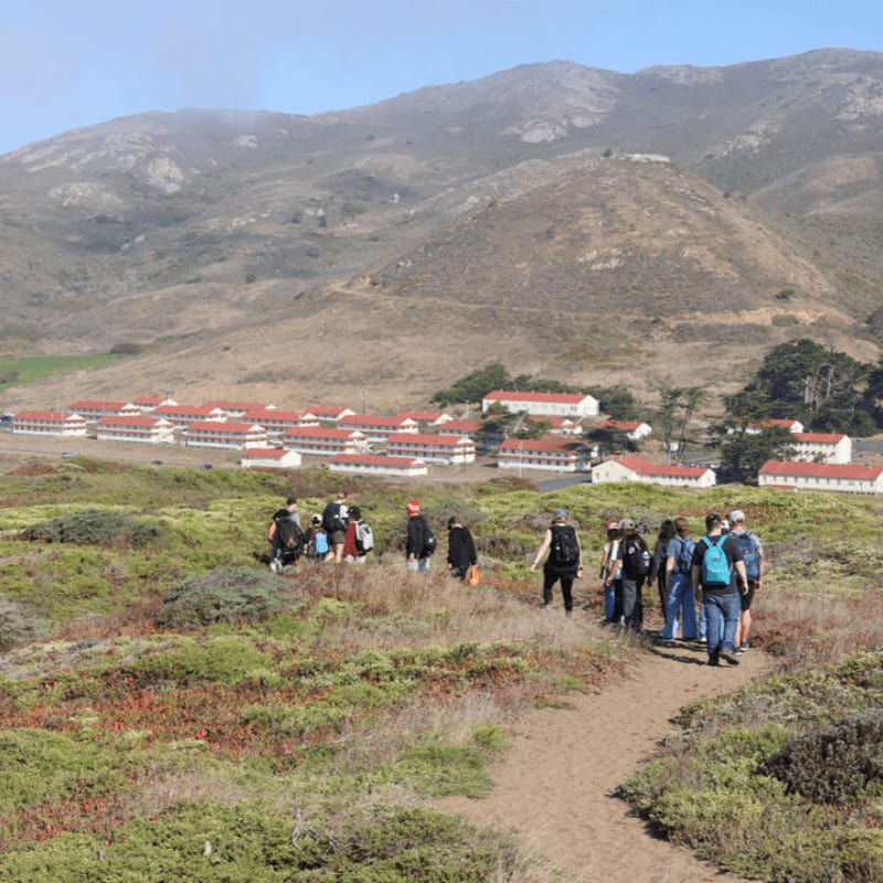 A group of hikers walks along a dirt path through scrubland toward a cluster of white buildings with red roofs set against a backdrop of large, brown mountains.