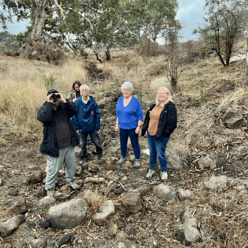Four people stand on a rocky path in the foreground, with a backdrop of dry grass and sparse trees under a cloudy sky.
