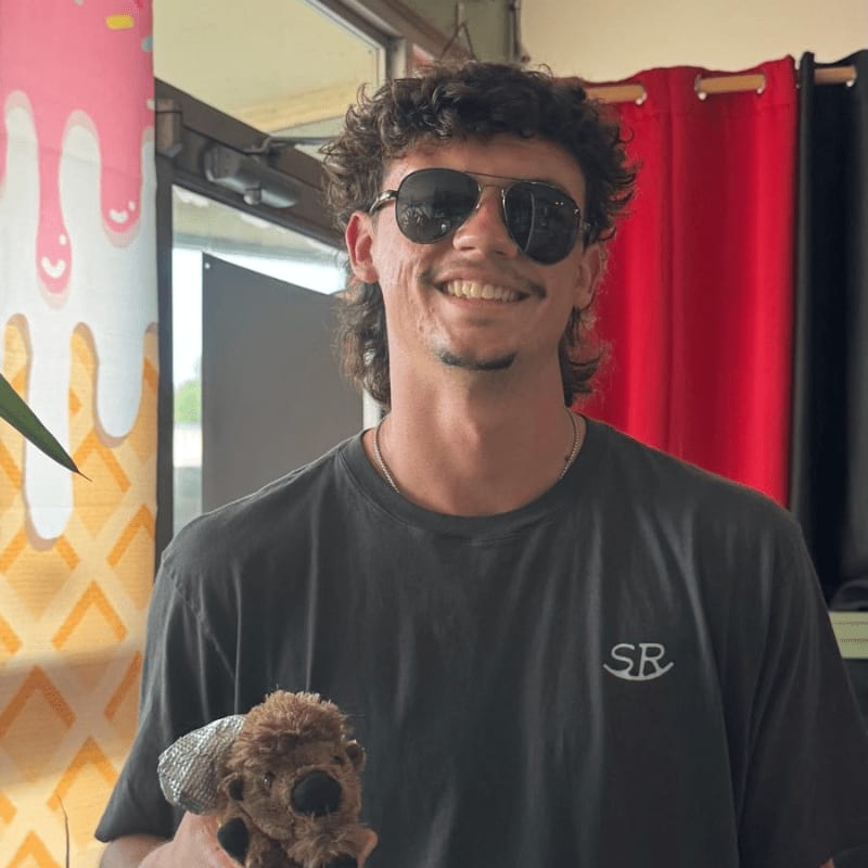 A smiling man wearing sunglasses and a black t-shirt holds a small teddy bear in front of a background featuring a red curtain and a colorful ice cream graphic.