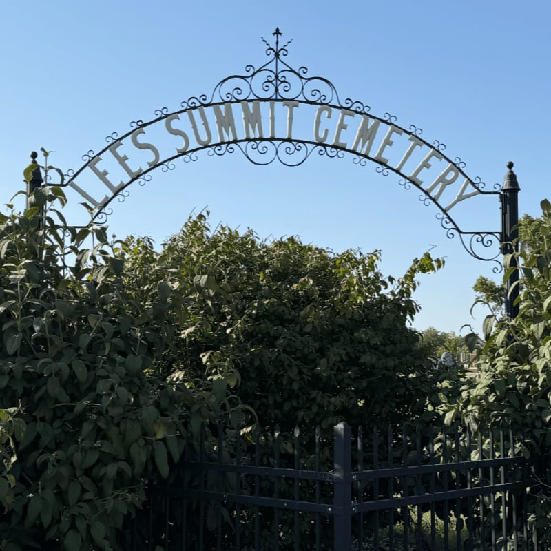 An ornate wrought-iron arch reading "LEES SUMMIT CEMETERY" stands above a black fence and lush green bushes against a clear blue sky.