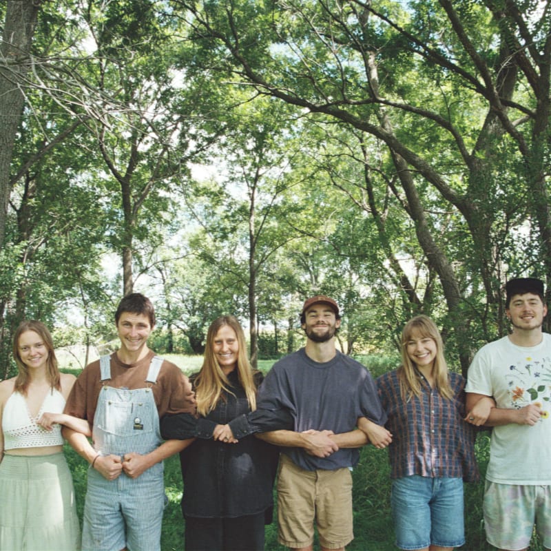 Six people stand side-by-side in the foreground against a background of lush green trees.
