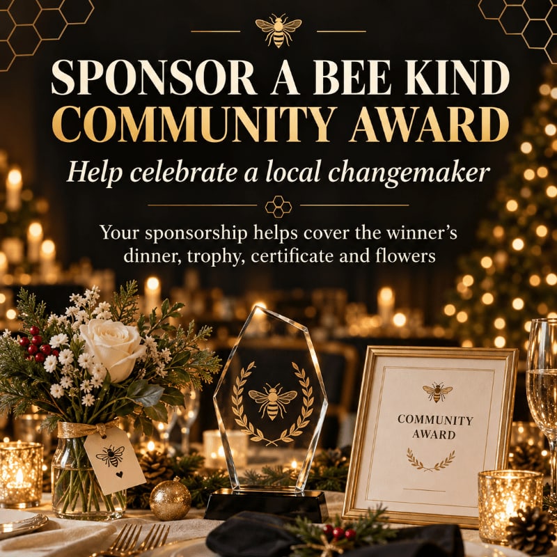 A crystal bee trophy, a framed award, and a floral bouquet sit on a dinner table against a blurred background of warm candlelight and festive lights.