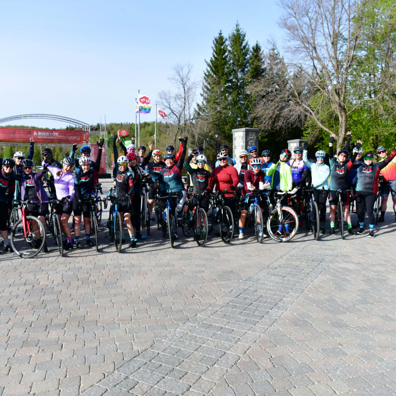 A group of cyclists in colorful gear pose with their bikes on a cobblestone plaza, with trees and a red archway in the background.