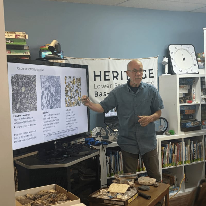 A man stands in the foreground next to a presentation screen about rock identification, with a bookshelf and a large clock in the background.