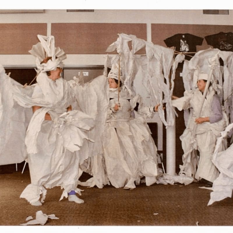 Three people wear voluminous white paper costumes in a room with neutral-toned walls and dark clothing hanging in the background.