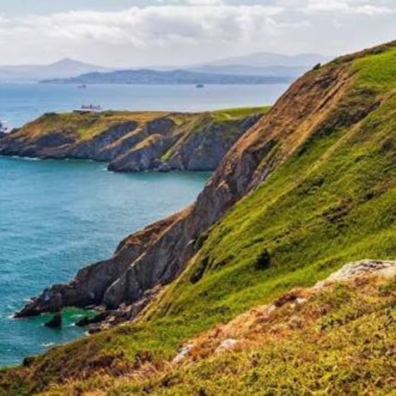 Lush green cliffs slope down to a turquoise sea, with distant mountains under a cloudy sky in the background.