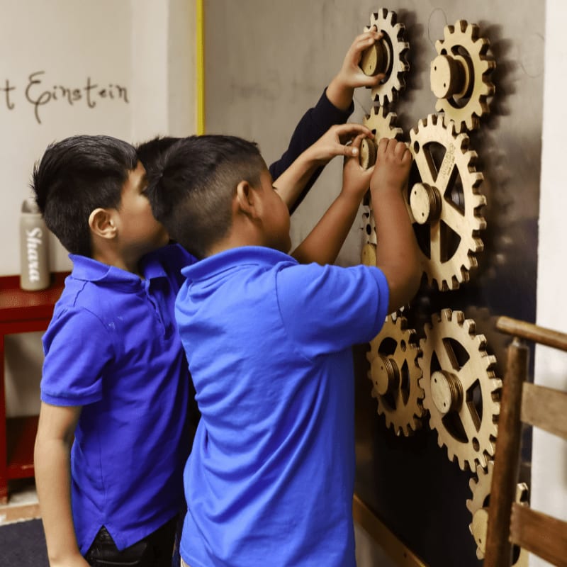 Three young boys in blue shirts interact with a wall of wooden gears in a room with a white wall.