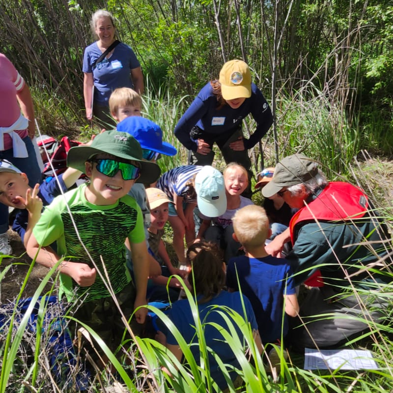 A group of children and adults gather in a grassy clearing surrounded by dense green trees and foliage.