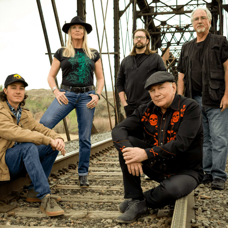 Five people in casual attire pose on a railroad track in front of a large steel bridge structure.