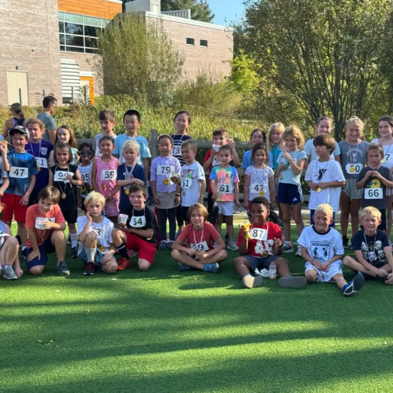 A group of young runners in racing bibs pose together on a green field in front of a modern building and lush greenery.