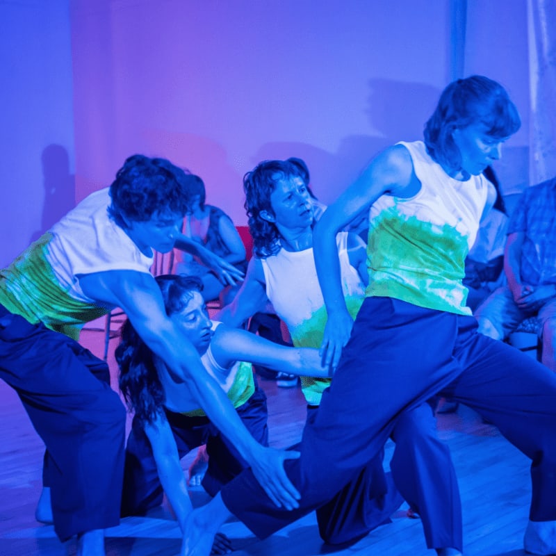 Several performers in white and green tops and dark pants engage in a dynamic dance sequence against a plain, blue-lit background.