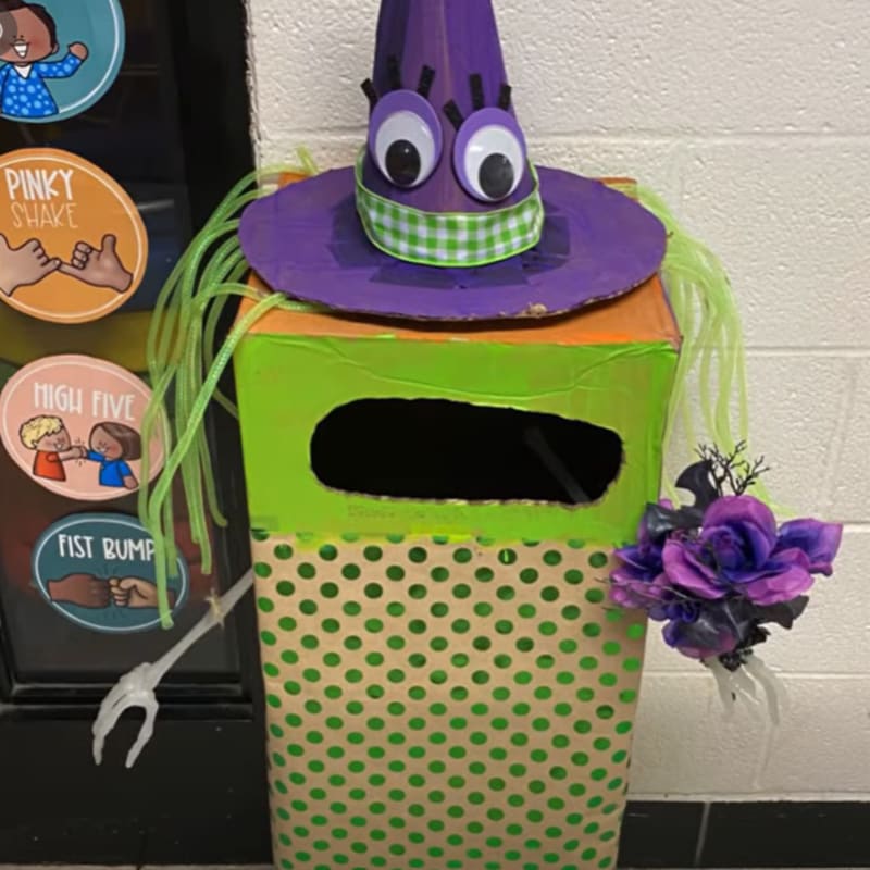 A cardboard witch craft with a purple hat and green polka dots stands against a white brick wall and a black door decorated with greeting circles.