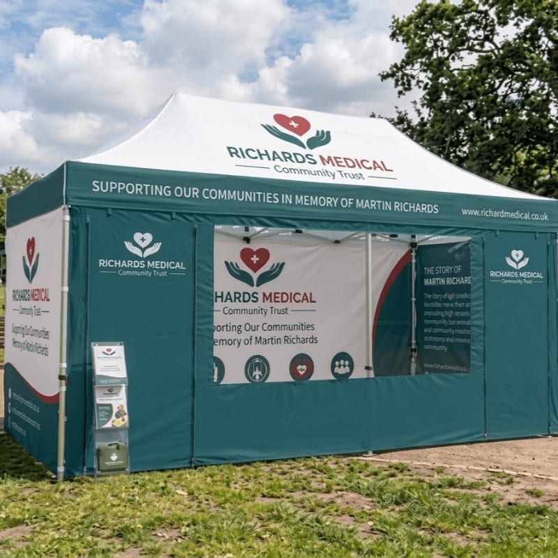 A branded Richards Medical Community Trust promotional tent stands on a grassy field against a backdrop of green trees and a cloudy blue sky.