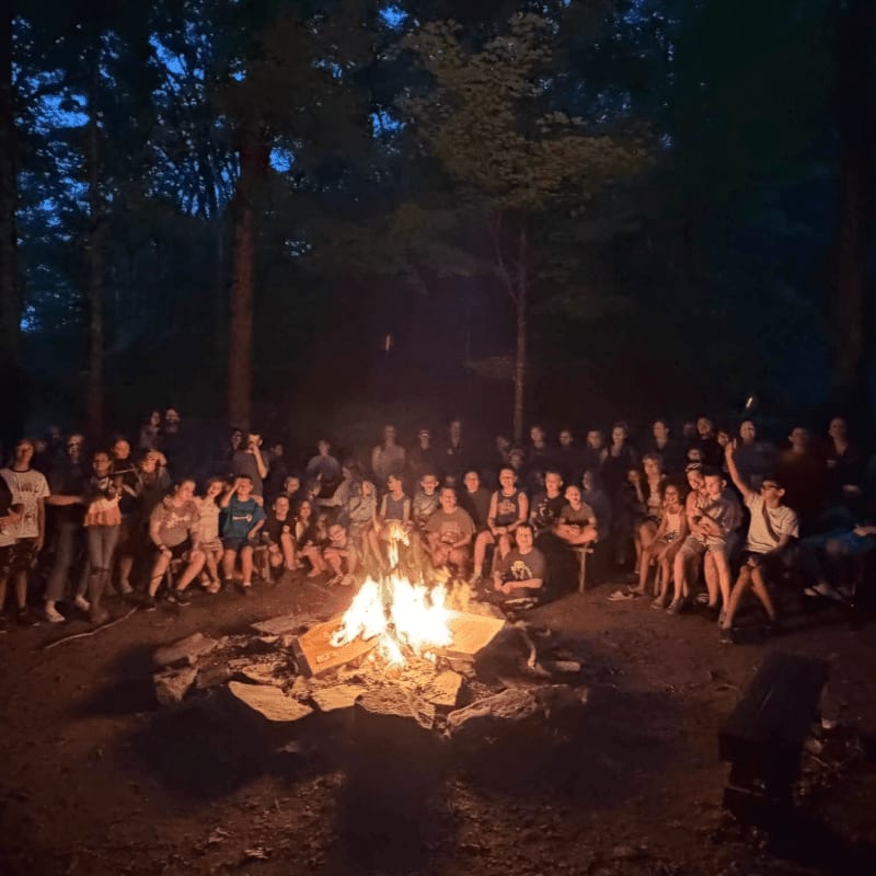 A group of people sit around a crackling campfire in a dark forest clearing.