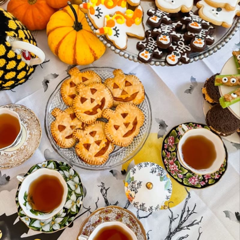 Pumpkin-shaped cookies and tea cups sit in the foreground on a white tablecloth, with pumpkins and a platter of themed treats in the background.
