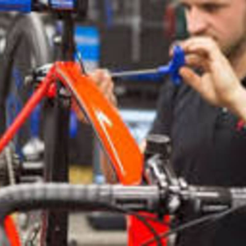 A mechanic in a workshop tightens a bolt on an orange bicycle frame, with blurred tools and equipment in the background.