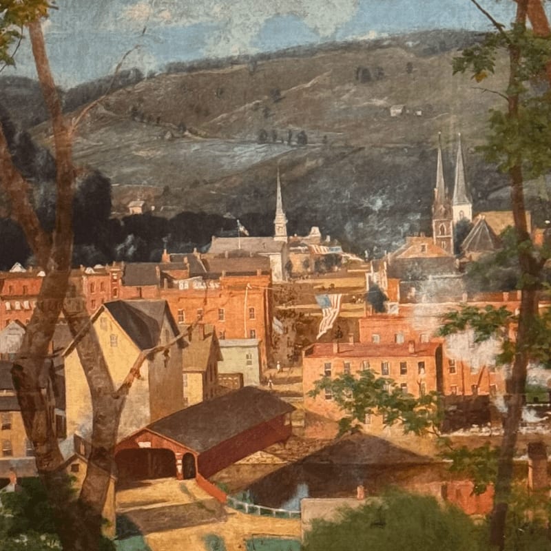 Brick buildings and a red covered bridge sit in the foreground, while a town with church spires rests at the base of a large, rolling hill in the background.