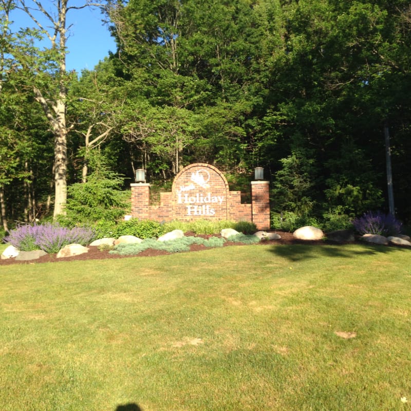 A brick sign for "Holiday Hills" sits behind a manicured lawn and flowerbed, backed by a dense forest of green trees.