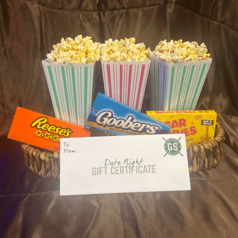 A date night gift basket containing popcorn boxes, candy, and a gift certificate sits against a draped brown fabric background.