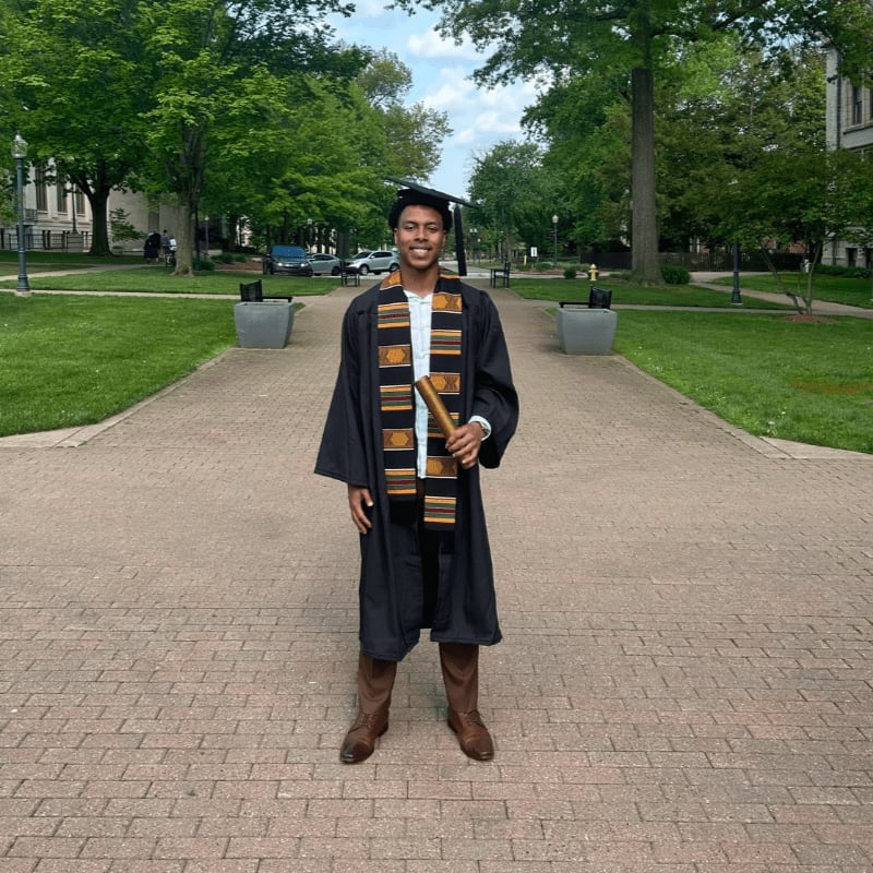 A graduate in a black gown and kente stole stands on a brick path surrounded by green grass and lush trees.