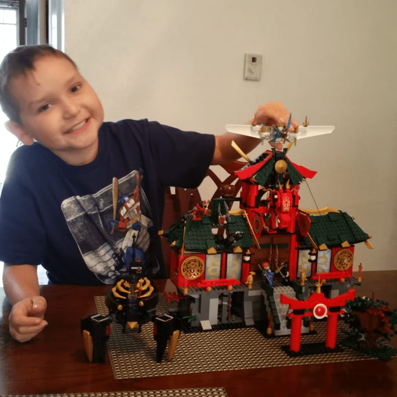 A smiling young boy in a blue shirt plays with a large, red and green LEGO temple set on a wooden table against a plain white wall.