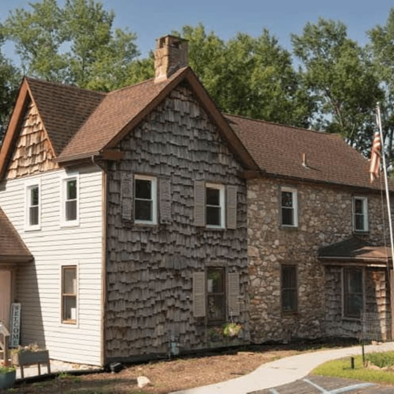 A rustic house with mixed wood shingle and stone siding stands before a backdrop of lush green trees.