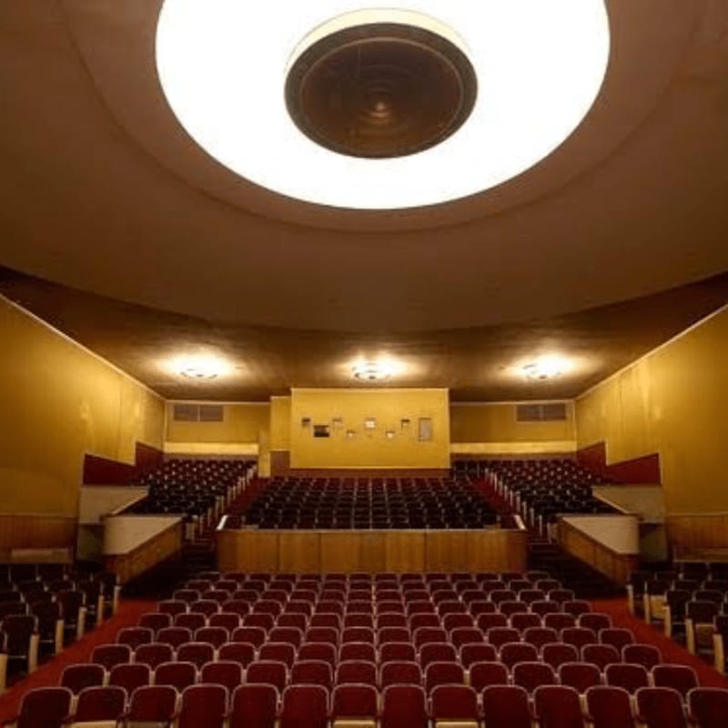 Rows of red theater seats fill the foreground of a large auditorium with yellow walls and a circular ceiling light in the background.