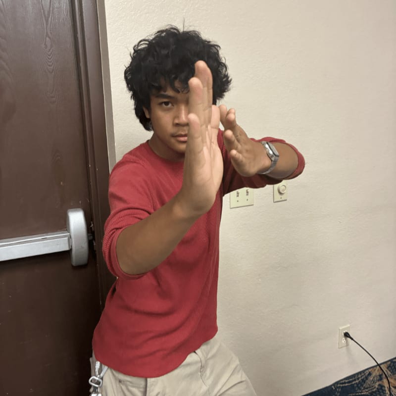 A young man in a red shirt holds his hands up in a defensive pose against a background of a white wall and a brown door.