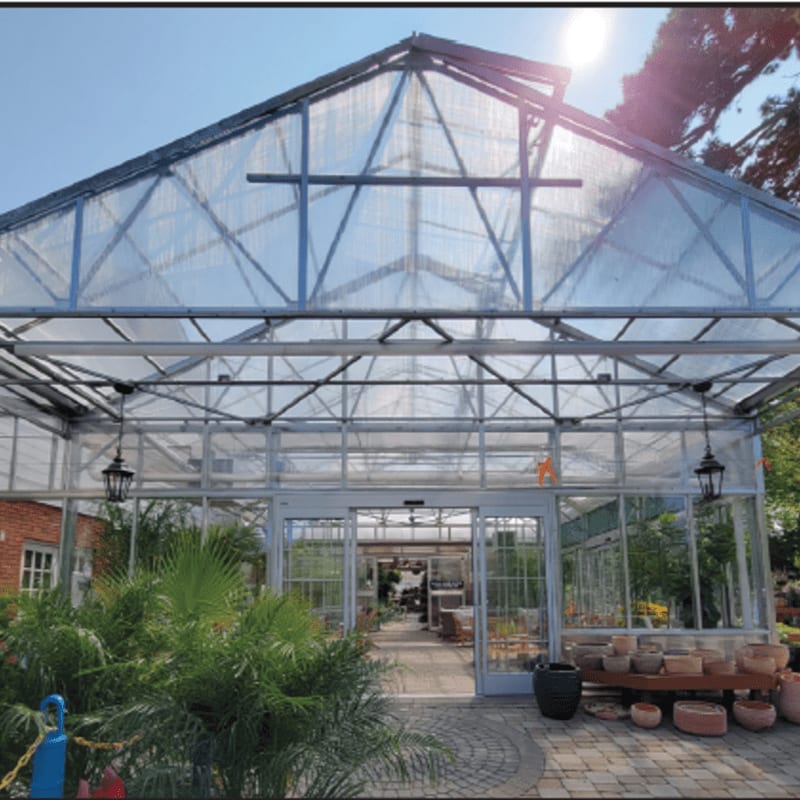 A large glass greenhouse stands in the background behind lush green plants and various potted ceramics in the foreground.