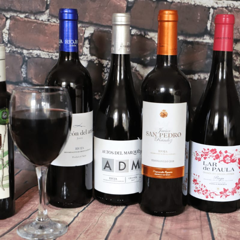 A glass of red wine stands in the foreground before a row of five wine bottles set against a brick wall background.