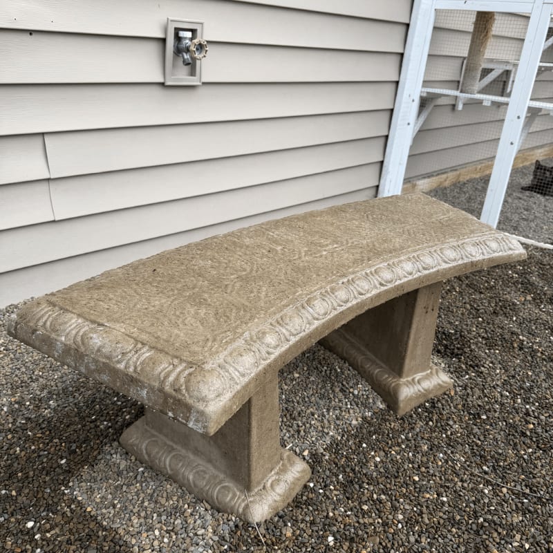 A weathered stone bench sits on a gravel surface in front of a light-colored sided wall and a white fence.