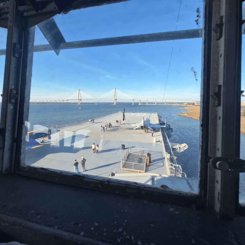 A view through a window frames the deck of an aircraft carrier with people on it, set against a backdrop of a wide river and a large cable-stayed bridge.