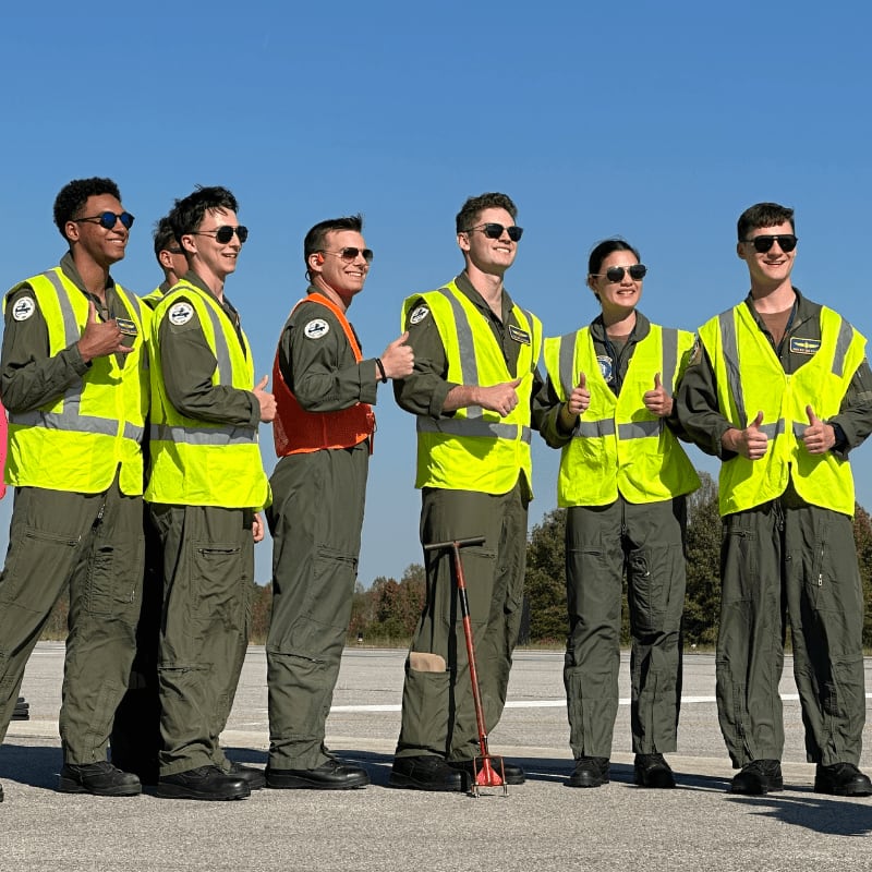 Six people in flight suits and high-visibility vests stand and give thumbs-up gestures on an airfield under a clear blue sky.
