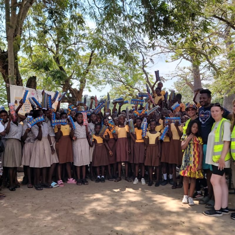 A group of school children and adults hold blue packages in the foreground, standing beneath the shade of large green trees in the background.