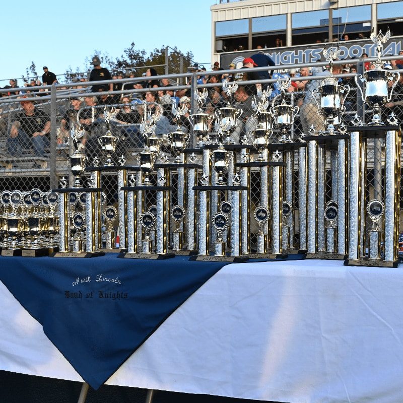 Numerous silver trophies stand on a table covered with a blue and white cloth, with a crowd of people and a stadium building in the background.