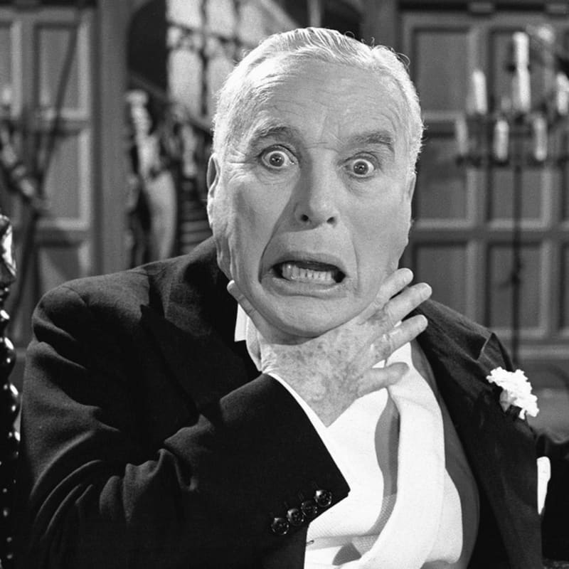 A wide-eyed man in a tuxedo clutches his neck in terror against a blurred interior background.