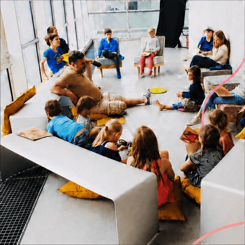 A group of children and adults sit and lounge in a bright, modern room with large windows.